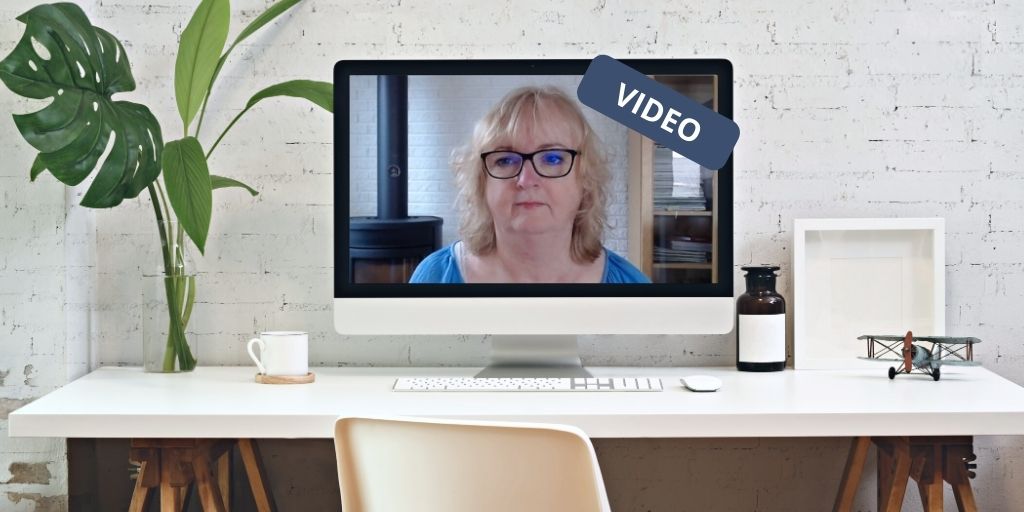 Computerskærm på skrivebord med billede af kvinde med briller. Ved siden af er der en plante og kaffekop. På den anden side en flaske, en lille flyvemaskine og en kasse.